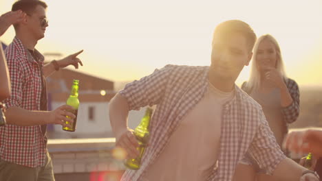 The-young-men-is-dancing-on-the-roof-with-his-friends-who-drinks-beer-on-the-party.-He-smiles-and-enjoys-the-time-in-shorts-and-a-blule-plaid-shirts-in-summer-evening.